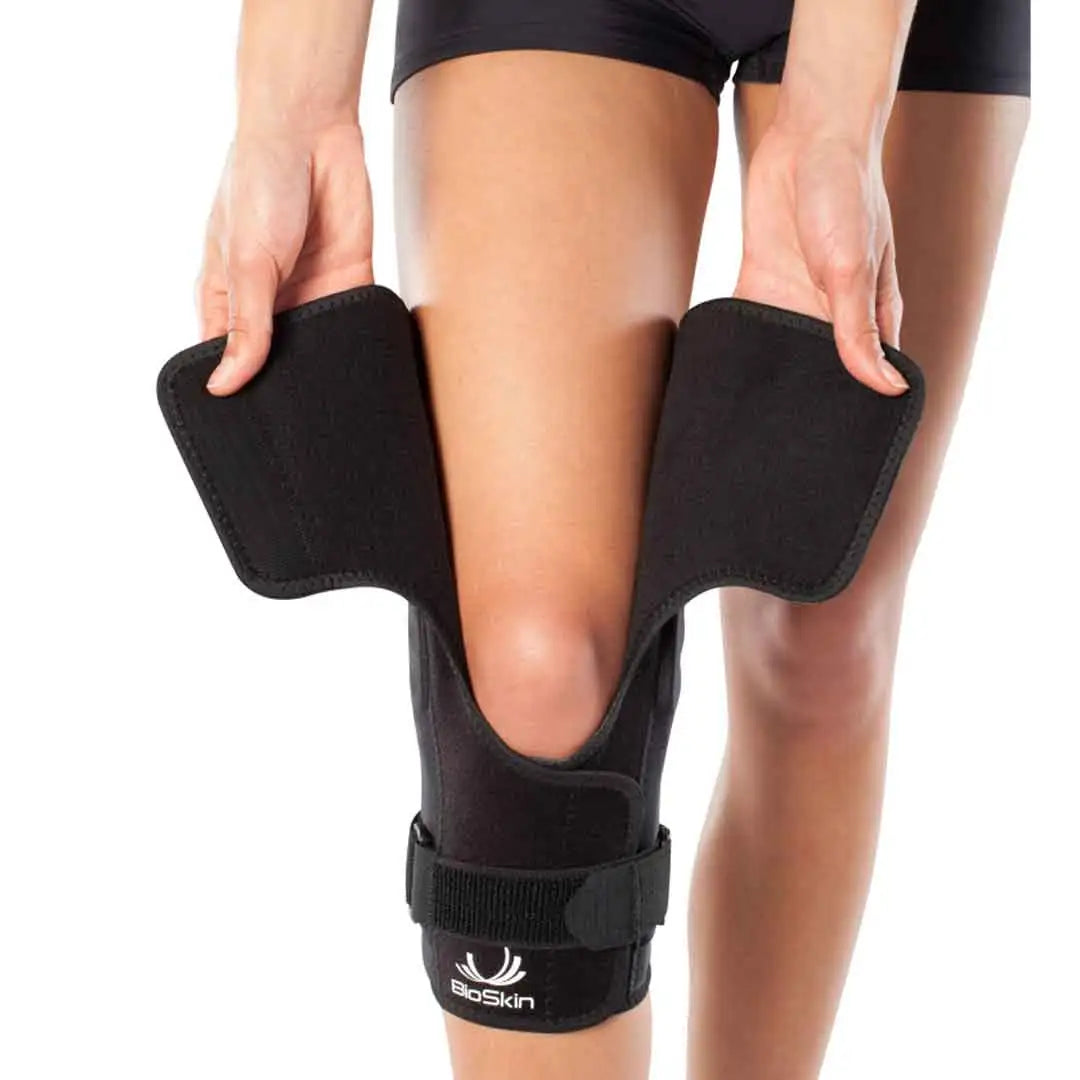 Das Bild zeigt die BioSkin Hinged Knee Sleeve Wraparound, eine stützende Kniebandage mit seitlichen Stäben zur Stabilisierung des Kniegelenks. Die Bandage besteht aus einem leichten, atmungsaktiven Material und ist so gestaltet, dass sie das Knie umschliesst und mit Klettverschlüssen individuell angepasst werden kann. 