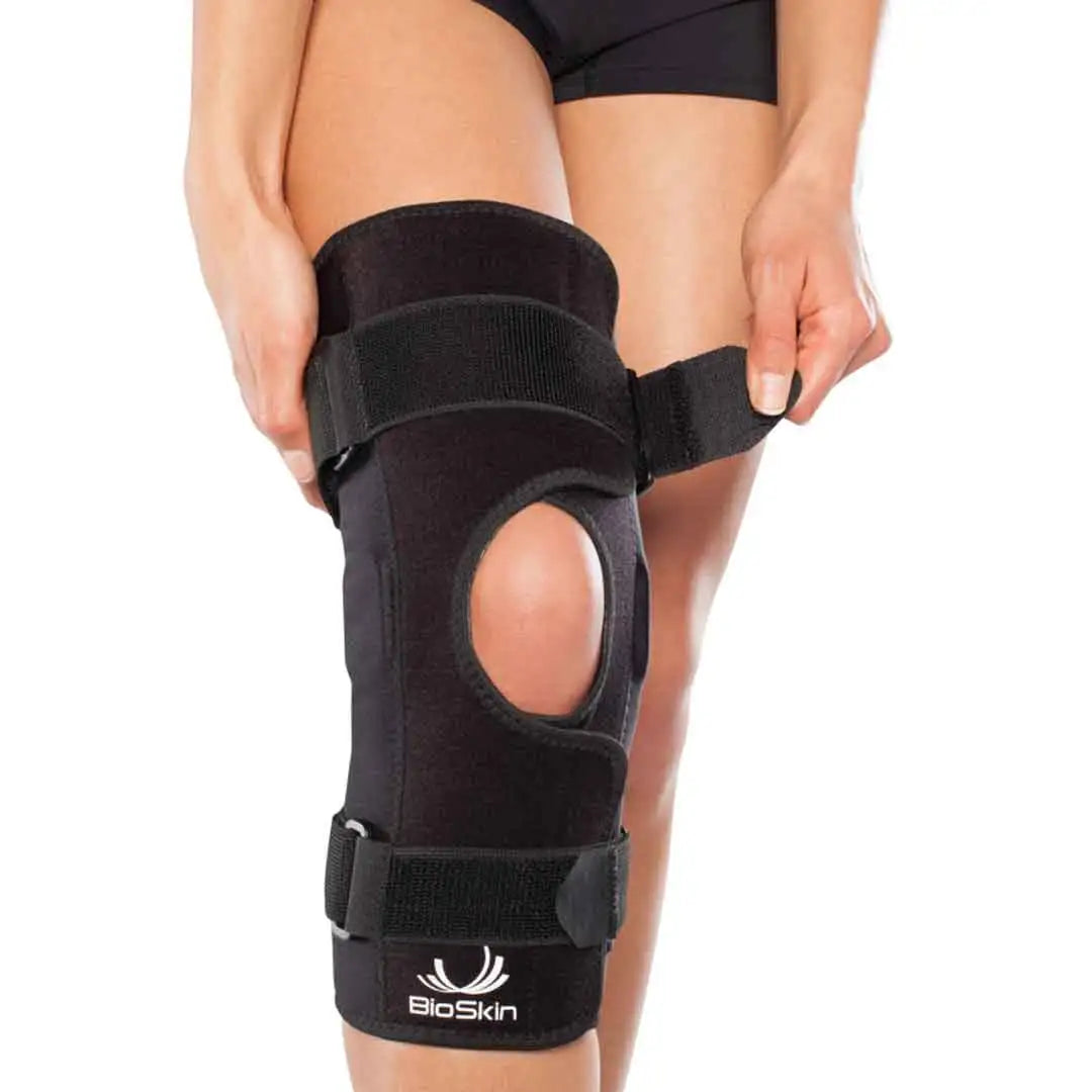 Das Bild zeigt die BioSkin Hinged Knee Sleeve Wraparound, eine stützende Kniebandage mit seitlichen Stäben zur Stabilisierung des Kniegelenks. Die Bandage besteht aus einem leichten, atmungsaktiven Material und ist so gestaltet, dass sie das Knie umschliesst und mit Klettverschlüssen individuell angepasst werden kann. 